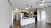 This single-floor residence is designed with an open-concept layout that seamlessly integrates the kitchen and living areas into one cohesive space. The kitchen serves as the focal point, anchored by a generously sized central island topped with light granite-style countertops, offering ample workspace and casual seating potential. Two pendant lights hang above the island, providing targeted task lighting that complements the flush ceiling fixture overhead.

The cabinetry throughout the kitchen is finished in a rich, dark espresso tone, creating a striking contrast against the lighter countertops and white walls. Stainless steel appliances, including a refrigerator and range with overhead microwave, add a modern, polished aesthetic to the cooking space. Additional cabinetry extends along the perimeter walls, providing substantial storage capacity.

The flooring throughout the space consists of wood-style planks in a warm, medium-toned finish, lending warmth and continuity across the open floor plan. Architectural details such as crown molding and columnar accents contribute a refined quality to the overall design. Multiple doorways visible from the main area suggest well-organized access to additional rooms. The overall ambiance is clean, modern, and functional.