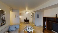 This single-floor space features an open-concept layout that seamlessly connects the living, dining, and kitchen areas. The living area is carpeted in warm beige tones and is anchored by a ceiling fan with a light fixture, providing both ventilation and overhead illumination. A sofa positioned near the wall and a small bookshelf along the right side offer comfortable and practical furnishings.

The dining area sits centrally within the open space, accommodating a compact round table with seating for two, making it well-suited for intimate dining. Just beyond, the kitchen is partially visible, featuring cabinetry and appliances, including a refrigerator, set against light-colored walls.

A walk-in closet with sliding doors is prominently featured along the back wall, offering generous storage space with visible shelving and hanging areas. An electrical panel is mounted nearby, housed neatly on the wall. The overall layout is efficient and well-organized, with neutral wall tones and carpeting contributing to a warm, understated ambiance throughout the space.