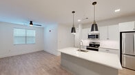 This single-floor residence showcases a clean, contemporary design characterized by bright white walls, light wood-look flooring, and an open, airy layout. The kitchen serves as the centerpiece of the main living area, featuring a large center island with a white quartz countertop and an integrated sink, complemented by sleek pendant lighting with matte black metal shades. White shaker-style cabinetry lines the walls, accented by a geometric patterned tile backsplash, and the kitchen is fully equipped with stainless steel appliances including a refrigerator, range, and microwave. Recessed lighting illuminates the space throughout.

The bedrooms offer a comfortable contrast with neutral carpet flooring and ceiling fans in each room. The primary bedroom suite provides generous storage with access to a large walk-in closet featuring built-in shelving, along with a dedicated laundry area nearby. Additional rooms offer ample space and natural light from blinds-fitted windows.

The bathroom is designed with a dual-sink vanity topped with white counters, framed mirrors above each sink, and a glass-enclosed walk-in shower with light tile walls. The consistent use of light tones, modern fixtures, and thoughtful storage solutions throughout creates a cohesive, functional, and well-appointed living environment.