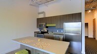 This single-floor space showcases a thoughtfully designed interior that blends modern aesthetics with industrial character. The kitchen is a standout feature, boasting a full suite of stainless steel appliances — including a refrigerator, electric range, built-in microwave, and dishwasher — paired with rich, dark wood-grain cabinetry that extends to the ceiling for generous storage. A pop of lime green accent on the backsplash adds a vibrant, contemporary touch, complementing the cool-toned palette throughout. The expansive kitchen island with a light neutral countertop provides ample prep and dining space, making it a central gathering point. Soaring ceilings amplify the sense of openness and volume, while exposed metal ductwork and a structural grid ceiling lend the space a distinctive industrial-loft character. The open layout promotes a seamless flow and an airy atmosphere. Clean lines, functional design, and carefully chosen accent details come together to create a space that is both visually engaging and highly practical for everyday living.