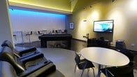 This single-floor entertainment lounge is thoughtfully designed to serve as a dynamic social and recreational hub. The space is defined by its bold, contemporary aesthetic, featuring yellow-painted walls accented with ambient lighting that creates an energetic yet welcoming atmosphere. Recessed cove lighting with blue-toned illumination highlights a textured feature wall in the background, adding a striking visual layer to the room.

At the heart of the space sits a sleek black pool table, surrounded by bar-height seating that encourages casual gathering. A wall-mounted flat-screen television is positioned for comfortable viewing from multiple vantage points, supported by an entertainment console with media equipment. Adjacent to the TV wall, a round white table with modern black chairs provides a designated area for conversation or tabletop activities.

Plush black reclining lounge chairs with built-in cup holders are arranged toward the front of the space, offering a comfortable spot to unwind. Track lighting along the ceiling supplements the ambient glow, ensuring the room is well-lit for various activities. The polished concrete-style flooring ties the design elements together, giving the space a clean, urban feel. Overall, this lounge delivers a versatile, stylish environment suited for entertainment and social interaction.