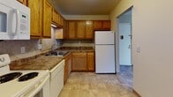 This single-floor unit offers a practical and comfortable layout spread across several distinct areas. The living room is generously sized, featuring wall-to-wall beige carpeting, neutral tan walls, and a ceiling fan overhead. A wall-mounted air conditioning unit provides supplemental cooling, while baseboard heaters run along the walls for warmth. A standout feature of the living room is a set of French doors that open to a private balcony, allowing natural light to pour in and offering outdoor views. A large closet with louvered bifold doors adds convenient storage directly off this main area.

The kitchen is equipped with warm oak cabinetry, dark granite-style countertops, a tile backsplash, and light tile flooring. White appliances including a refrigerator, dishwasher, and range are included. The kitchen connects to an entryway with hard flooring near the unit's front door.

The bedrooms are carpeted and finished with the same neutral tones found throughout. Each room benefits from natural light via double-hung windows fitted with blinds. One bedroom includes a closet and baseboard heating. The bathroom features a full-sized white soaking tub and shower combination with a built-in corner shelf and chrome fixtures, maintaining a clean, functional aesthetic throughout.