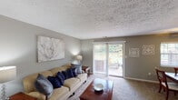 This single-floor residence offers a thoughtfully arranged layout that balances comfort and functionality throughout. The living room serves as the heart of the home, featuring soft wall-to-wall carpeting, neutral gray walls, and a textured ceiling. A sliding glass door floods the space with natural light and opens to an outdoor area, providing pleasant views of greenery beyond. The room flows seamlessly into the dining area, which is highlighted by a ceiling fan with integrated lighting and direct sightlines into the kitchen — ideal for entertaining. The galley-style kitchen is efficiently designed with ample white cabinetry on both sides, a double-basin sink, a full suite of white appliances including a range, microwave, dishwasher, and refrigerator, and durable tile flooring with a rich, earthy tone. The bedroom is a restful retreat, showcasing a bold accent wall in deep navy blue, plush carpeting, a natural light source through a window with blinds, and a generously sized open closet for storage. The bathroom completes the space with sage green walls, tile flooring, a tub-shower combination, and a well-lit vanity, creating a calm and functional environment throughout.