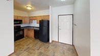 This single-floor unit in Building 1 offers a practical and well-organized layout across several distinct living spaces. The living area features beige wall-to-wall carpeting, a textured ceiling, and a sliding glass door with vertical blinds that allows natural light to filter in. A baseboard heating unit runs along the exterior wall, and the space flows seamlessly into the kitchen and dining area via stone-look tile flooring.

The kitchen is equipped with warm-toned wooden cabinetry, dark countertops, and a full suite of black appliances, including a refrigerator, dishwasher, electric range, and over-the-range microwave. A stainless sink is positioned beneath upper cabinetry, and a dedicated laundry closet just off the kitchen houses a stacked washer and dryer unit, providing added convenience.

The bedrooms are carpeted and finished with neutral white walls and wood-toned baseboards. Each room includes a window with horizontal blinds and baseboard heating, creating a functional and comfortable environment. The full bathroom features stone-look tile flooring, a single-sink vanity with wooden cabinetry, a wall-mounted mirror with lighting, and a tub-shower combination.