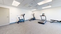 This fitness center in Phoenix, AZ, is a single-room exercise facility designed to provide a practical and functional workout environment. The space is efficiently organized to accommodate a variety of fitness equipment within a compact footprint, making it suitable for focused individual training sessions.

The exercise room features a drop-panel ceiling with recessed fluorescent lighting that evenly illuminates the space. The flooring throughout consists of durable, speckled low-pile carpet, providing cushioning and traction suitable for fitness activities. Neutral white walls with a gray base trim create a clean, minimal aesthetic throughout the room.

The facility is equipped with multiple cardio machines, including a treadmill, elliptical trainer, and stationary bike, positioned to allow comfortable usage. A multi-function cable weight machine serves as the strength-training centerpiece, complemented by a rack of free weights and a wall-mounted exercise mat for stretching or floor work. A large window on one wall admits natural light and provides views of the surrounding Phoenix cityscape, adding an open feel to the otherwise interior-focused space. Access is provided through a framed door with a window panel, connecting to adjacent areas of the building.