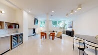 Located on Santa Monica Boulevard in Los Angeles, this thoughtfully designed single-floor amenity space offers a bright, contemporary environment ideal for both relaxation and collaboration. The space is anchored by polished terrazzo flooring throughout, lending a clean and cohesive aesthetic across all areas.

The main lounge area is an inviting, open layout featuring a curved cream sectional sofa, bold orange accent chairs, and a wall-mounted fireplace with an integrated TV. Structural columns define zones within the open floor plan without disrupting the sense of spaciousness. Large windows along the perimeter flood the interior with natural light and offer views of the tree-lined streetscape, enhancing the airy ambiance. Curated artwork and sculptural décor add warmth and artistic character to the neutral white walls.

A dedicated conference room provides a more private setting, furnished with a dark oval table, white upholstered chairs, and a large-scale abstract painting. Greenery and recessed lighting contribute to a focused yet comfortable atmosphere.

The kitchenette is well-appointed with light wood cabinetry, white quartz countertops, open shelving, a stainless steel sink, a warming drawer, and dual wine/beverage refrigerators — offering full amenity support for gatherings or day-to-day use.