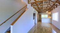 This unique multi-floor space makes an immediate impression with its dramatic exposed timber roof trusses, which span the upper level and lend the interior a warm, structural character rarely found in standard builds. The open upper floor showcases light-toned hardwood flooring that complements the natural wood tones of the overhead framework, creating an airy, loft-like atmosphere filled with natural light from a generous window at the far end. A wood-railed staircase with clean lines connects the levels, contributing to the cohesive use of natural wood throughout. A built-in closet with open shelving is thoughtfully integrated into the upper floor's layout, providing practical storage without disrupting the open feel of the space. The bathroom, accessible via a wood-framed doorway, features classic small-tile flooring, a toilet, and a vanity with cabinet storage beneath — a straightforward and functional design consistent with the overall character of the property. White walls throughout all levels keep the aesthetic bright and neutral, allowing the architectural details — particularly the exposed trusses — to remain the focal point. Spanning three floors, this space offers a versatile and characterful environment well-suited for a variety of uses.