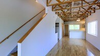 This unique multi-floor space makes an immediate impression with its dramatic exposed timber roof trusses, which span the upper level and lend the interior a warm, structural character rarely found in standard builds. The open upper floor showcases light-toned hardwood flooring that complements the natural wood tones of the overhead framework, creating an airy, loft-like atmosphere filled with natural light from a generous window at the far end. A wood-railed staircase with clean lines connects the levels, contributing to the cohesive use of natural wood throughout. A built-in closet with open shelving is thoughtfully integrated into the upper floor's layout, providing practical storage without disrupting the open feel of the space. The bathroom, accessible via a wood-framed doorway, features classic small-tile flooring, a toilet, and a vanity with cabinet storage beneath — a straightforward and functional design consistent with the overall character of the property. White walls throughout all levels keep the aesthetic bright and neutral, allowing the architectural details — particularly the exposed trusses — to remain the focal point. Spanning three floors, this space offers a versatile and characterful environment well-suited for a variety of uses.