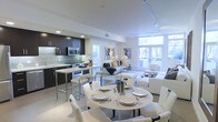 This single-floor space showcases a thoughtfully designed open-plan layout that seamlessly integrates the kitchen, dining, and living areas into one cohesive environment. The kitchen features dark espresso cabinetry contrasted against light countertops and a breakfast bar with chrome-legged stools, offering both functionality and style. Stainless steel appliances, including a built-in microwave and range, complement the contemporary aesthetic.

The dining area is anchored by a round table with a studded base, surrounded by cream upholstered chairs, creating an inviting setting for meals. Natural light floods the space through large floor-to-ceiling windows and French doors, which provide outdoor views and enhance the airy, open atmosphere throughout.

The living area features a neutral palette of white and grey furnishings, including plush sofas and a lounge chair, arranged to encourage relaxed conversation. Track lighting runs along the ceiling, supplementing the abundant natural light and allowing for flexible illumination. The smooth, light-toned flooring ties the entire space together, reinforcing the clean, modern sensibility that defines this well-appointed interior.
