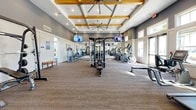 Located at 725 Flagler Crossing Drive in St. Augustine, FL, this fitness center offers a generously sized, single-floor layout divided into two dedicated exercise rooms. The main exercise area impresses with soaring vaulted ceilings accented by exposed wooden beams and oversized ceiling fans, creating an open, airy atmosphere. Neutral gray walls, plank-style flooring, and plush carpet tiles provide a modern, clean aesthetic throughout. Floor-to-ceiling windows flood the space with abundant natural light and offer views of the lush outdoor surroundings, including palm trees and the surrounding community. The primary room is lined with full-length mirrors and equipped with a comprehensive array of strength machines, cable systems, a power rack with bench, treadmills, ellipticals, and rowing machines, complemented by wall-mounted televisions for entertainment. A secondary room serves as a functional training zone, featuring TRX suspension systems, kettlebells, medicine balls, stability balls, and a plyo box, along with stationary bikes and additional cardio equipment. Motivational wall art and thoughtful décor complete the inviting environment, making this fitness center a well-appointed destination for a full range of workouts.