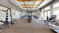 Located at 725 Flagler Crossing Drive in St. Augustine, FL, this fitness center offers a generously sized, single-floor layout divided into two dedicated exercise rooms. The main exercise area impresses with soaring vaulted ceilings accented by exposed wooden beams and oversized ceiling fans, creating an open, airy atmosphere. Neutral gray walls, plank-style flooring, and plush carpet tiles provide a modern, clean aesthetic throughout. Floor-to-ceiling windows flood the space with abundant natural light and offer views of the lush outdoor surroundings, including palm trees and the surrounding community. The primary room is lined with full-length mirrors and equipped with a comprehensive array of strength machines, cable systems, a power rack with bench, treadmills, ellipticals, and rowing machines, complemented by wall-mounted televisions for entertainment. A secondary room serves as a functional training zone, featuring TRX suspension systems, kettlebells, medicine balls, stability balls, and a plyo box, along with stationary bikes and additional cardio equipment. Motivational wall art and thoughtful décor complete the inviting environment, making this fitness center a well-appointed destination for a full range of workouts.