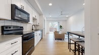 This well-appointed single-floor unit offers a thoughtfully designed open-concept layout that seamlessly connects the kitchen and living areas. The kitchen is outfitted with crisp white shaker cabinetry, matte black hardware, a classic white subway tile backsplash, grey countertops, and a full suite of black appliances including a range, over-the-range microwave, and dishwasher. Light hardwood flooring runs throughout the main living space, enhancing the bright, airy atmosphere created by recessed lighting and a modern ceiling fan overhead. The living area is generously proportioned, anchored by large sliding glass doors that invite natural light and provide access to an outdoor area with views of surrounding greenery. A dedicated office features plush carpeting, a wall-mounted mini-split HVAC unit, and a window overlooking the street, making it a comfortable and functional workspace. The bathroom is finished with large-format tile flooring, white shaker vanity cabinetry with grey countertops, a soaking tub with bold black tile surround, and a sleek barn-style sliding door that connects directly to the in-unit laundry room. The laundry room houses a full-size front-loading washer and dryer alongside open shelving for convenient storage. Clean white walls and consistent modern finishes unify the space throughout.