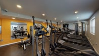 This fitness center is a comprehensively outfitted exercise space designed to support a full range of workout routines. The room features durable rubber flooring throughout, providing a safe and functional surface for all types of training activity. Recessed lighting illuminates the space evenly from above, while natural light filters in through multiple windows fitted with blinds along one wall, contributing to an energizing atmosphere.

The cardio section is well-stocked with a row of SportsArt treadmills and elliptical cross-trainers, offering ample capacity for aerobic exercise. Complementing these are several strength training stations, including a multi-functional cable machine and a plate-loaded machine, catering to a variety of resistance training needs. A dedicated free weights area includes a full dumbbell rack with a range of weights and an adjustable workout bench for versatile strength exercises.

A large wall-mounted flat-screen television is positioned prominently for entertainment during workouts. Accent walls painted in a bold yellow add a dynamic visual contrast to the neutral gray tones of the ceiling and surrounding walls. Wall mirrors are strategically placed to enhance the sense of space and allow users to monitor their form. The layout is thoughtfully organized to maximize usable floor space while keeping different training zones clearly defined.
