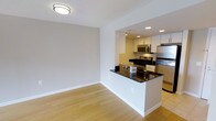 This single-floor space showcases a clean, contemporary design with an open-concept layout that seamlessly connects the living area to the kitchen. The living space is finished with light hardwood flooring that extends throughout, complementing the neutral gray walls and white baseboards. Recessed lighting in the kitchen, paired with a flush-mount ceiling fixture in the living area, ensures consistent illumination across both zones. The kitchen is defined by a peninsula with a dark granite countertop that doubles as a breakfast bar and visual divider between the two spaces. White shaker-style cabinetry lines the kitchen walls, providing ample storage, while stainless steel appliances — including a gas range, microwave, and refrigerator — add a modern, polished finish. The contrasting dark countertops against the white cabinetry create a striking visual balance. The open layout offers flexibility in furniture arrangement, and the overall design aesthetic is cohesive and functional, with an emphasis on clean lines and a neutral color palette that lends itself to a variety of interior styles.