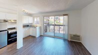 This single-floor unit offers a practical and well-organized layout with a comfortable, functional design throughout. The open living area is highlighted by dark hardwood-style flooring and neutral white walls, creating a clean and versatile space. A textured ceiling adds subtle character, while a sliding glass door floods the room with natural light and provides direct access to a private balcony overlooking a landscaped outdoor area with palm trees and neighboring buildings visible in the distance.

The kitchen is efficiently designed with ample white cabinetry above and below the countertops, offering generous storage space. Light-toned laminate counters provide a practical workspace, and the kitchen is equipped with an electric range, an overhead ventilation hood, a stainless steel double sink, and a countertop microwave. The kitchen opens toward the living area, promoting a connected and social atmosphere.

The bathroom features a clean, all-white walk-in shower stall with built-in shelving on both sides and chrome fixtures, alongside standard toilet facilities. The bright, minimalist design of the bathroom contributes to the overall fresh and well-maintained feel of the space.

With its combination of functional spaces, natural light, private outdoor access, and cohesive neutral finishes, this unit presents a straightforward and livable layout suited to a variety of needs.