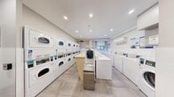 This shared laundry facility is a well-organized, single-floor space designed to accommodate high-volume laundry needs efficiently. The room features a clean, modern aesthetic with white tile wainscoting along the lower walls, a subtle textured wallcovering on the upper walls, and large-format light grey floor tiles that lend a bright, orderly appearance throughout.

Along one wall, a row of commercial-grade Speed Queen top-load washers is lined up in sequence, offering multiple units for simultaneous use. Opposite, a bank of stacked commercial dryers lines the wall, maximizing capacity within the footprint of the space. A central wooden folding counter provides a convenient surface for sorting and folding laundry, complemented by a stainless steel waste bin nearby.

A dedicated payment kiosk station is positioned near the entrance, allowing for card or app-based transactions, reflecting a modern, cashless laundry management system. Informational signage is posted throughout, including references to a smart laundry monitoring portal. A wall-mounted split air conditioning unit ensures year-round climate comfort.

Natural light filters in through a window at the far end of the room, while recessed ceiling lights provide consistent, even illumination across the entire space. The entrance features a solid-panel door alongside a windowed access door, contributing to a secure and well-ventilated environment.