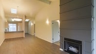This single-floor space is thoughtfully designed with an emphasis on architectural character and an open, airy atmosphere. The main living area immediately draws attention with its soaring vaulted ceiling, accented by exposed wooden beams that add warmth and a rustic yet refined aesthetic. A skylight positioned along the roofline allows natural light to cascade into the space, complementing the warm-toned hardwood flooring that runs throughout.

A standout feature of the living area is the bold, floor-to-ceiling shiplap fireplace surround finished in a deep charcoal-gray tone, creating a striking focal point that anchors the room. The fireplace insert is set within a clean-lined surround that balances modern design sensibilities with a cozy, inviting feel.

Wall sconces provide soft, ambient lighting along the hallway and adjoining areas, enhancing the welcoming atmosphere. The open layout connects the main living space to additional rooms accessed through crisp white paneled doors, suggesting a practical and well-organized floor plan. Neutral wall tones in soft beige and warm gray keep the palette cohesive and versatile throughout. Overall, the space combines vaulted architecture, natural materials, and carefully chosen design details to create a comfortable and visually engaging environment.