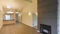 This single-floor space is thoughtfully designed with an emphasis on architectural character and an open, airy atmosphere. The main living area immediately draws attention with its soaring vaulted ceiling, accented by exposed wooden beams that add warmth and a rustic yet refined aesthetic. A skylight positioned along the roofline allows natural light to cascade into the space, complementing the warm-toned hardwood flooring that runs throughout.

A standout feature of the living area is the bold, floor-to-ceiling shiplap fireplace surround finished in a deep charcoal-gray tone, creating a striking focal point that anchors the room. The fireplace insert is set within a clean-lined surround that balances modern design sensibilities with a cozy, inviting feel.

Wall sconces provide soft, ambient lighting along the hallway and adjoining areas, enhancing the welcoming atmosphere. The open layout connects the main living space to additional rooms accessed through crisp white paneled doors, suggesting a practical and well-organized floor plan. Neutral wall tones in soft beige and warm gray keep the palette cohesive and versatile throughout. Overall, the space combines vaulted architecture, natural materials, and carefully chosen design details to create a comfortable and visually engaging environment.