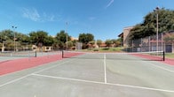 The tennis courts area is a well-appointed outdoor recreational facility comprising multiple hard-surface courts arranged side by side. Each court features a distinctive dual-tone surface with gray playing areas and terracotta-red boundary zones, all clearly marked with crisp white lines and fitted with regulation-height nets. Tall lighting poles are positioned throughout the complex, ensuring the courts remain usable during evening hours.

The courts are fully enclosed by chain-link fencing, providing both safety and containment during play. Mature trees line the perimeter, offering partial shade and a pleasant natural backdrop. The surrounding grounds are well-maintained, with neatly manicured green lawns, concrete walkways, and young ornamental trees that add to the landscaped aesthetic.

The facility is situated adjacent to multi-story residential buildings constructed in a warm, stucco-finished architectural style with earthy tan tones. Curved pathways connect the courts to nearby buildings, and globe-style lampposts along the walkways enhance both visibility and ambiance. The open-sky setting under clear blue skies contributes to an inviting, active outdoor environment ideal for recreational and competitive tennis play.
