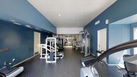 This fitness center offers a dedicated space for a comprehensive workout experience. The interior is defined by striking deep blue accent walls paired with crisp white ceilings, creating an energizing and focused atmosphere. Recessed lighting throughout ensures the space is well-illuminated at all times.

The gym floor is outfitted with durable rubber flooring, providing both safety and comfort during exercise. A variety of strength training equipment is prominently featured, including a Paramount multi-function cable machine and additional cable/pulley systems, catering to a full range of resistance training exercises. Cardio equipment, including a treadmill and a stationary bike, is also available for aerobic workouts.

Toward the back of the space, glass-panel doors allow natural light to filter in and provide views to an adjacent area, adding a sense of openness. The layout is thoughtfully organized, with equipment spaced to allow for comfortable movement between stations. Ventilation is maintained through visible wall vents, and the overall condition of the facility appears well-maintained. This fitness center serves as a practical and fully functional amenity designed to support a wide range of fitness goals.