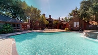 The Main Pool area is a well-appointed outdoor amenity designed for relaxation and recreation. At its center sits a generously sized freeform swimming pool with crystal-clear blue water, bordered by classic brick paver decking that adds a warm, traditional aesthetic. Depth markers along the pool's edge indicate a gradual transition from shallow to deeper waters, making it accessible for a range of swimmers. Attached to the pool is a raised spa, accented with brick surround detailing and featuring water jets, providing a dedicated space for hydrotherapy and leisure.

A covered gazebo with a pitched roof and sturdy columns offers shaded seating overlooking the pool, ideal for enjoying the outdoors without direct sun exposure. Multiple lounge chairs are arranged throughout the expansive patio, providing ample space for sunbathing and socializing. A separate poolside cabana-style building with large windows and French doors serves as a functional support structure for the amenity area. A gas grill is also available on the patio for outdoor cooking.

The entire space is framed by mature trees and lush greenery, creating a private, resort-like atmosphere. The surrounding multi-story residential buildings with brick detailing and multiple chimneys provide an attractive architectural backdrop under open blue skies.