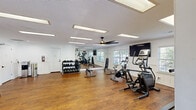 Located at 201 NC-54 W in Carrboro, North Carolina, this fitness center offers a comprehensive exercise environment within a single, open-plan room. The space features warm hardwood-style flooring throughout, crisp white walls with classic wainscoting detail, and a textured ceiling equipped with recessed panel lighting that ensures the room is consistently and brightly lit. A ceiling fan provides additional airflow and comfort during workouts.

The exercise room is thoughtfully organized into distinct functional zones. A cardio area houses a treadmill, elliptical trainer, stationary bikes, and a stair climber, with flat-screen TVs mounted on the walls for entertainment. A strength training section includes a multi-tiered dumbbell rack with a full range of free weights, accompanied by adjustable workout benches. A dedicated functional training zone features a well-stocked equipment rack holding TRX suspension trainers, resistance bands, medicine balls, kettlebells, stability balls, and exercise mats, along with slam balls for high-intensity training.

Large windows along the walls invite natural light and outdoor views, enhancing the open, airy atmosphere. A full-length mirror expands the sense of space and allows for form-checking during exercise. Vibrant, energetic wall murals add personality and motivation to the environment. A water dispenser is conveniently available for hydration. The overall ambiance is clean, energetic, and welcoming.