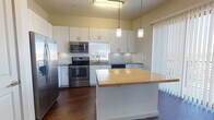 This well-designed single-floor residence offers a thoughtfully laid-out interior with a clean, contemporary aesthetic throughout. The kitchen serves as a standout feature, showcasing crisp white cabinetry, a classic subway tile backsplash, granite countertops, and a full suite of stainless steel appliances including a refrigerator, range, and microwave. A central island with a warm butcher block top provides additional prep space and casual seating potential, while pendant lighting adds a stylish, functional touch. A sliding glass door with vertical blinds floods the kitchen and adjacent areas with natural light and offers views to the exterior. Rich, dark hardwood flooring flows through the main living areas and hallways, lending warmth and continuity to the space. The bedrooms feature plush carpeting and large windows with horizontal blinds, allowing for natural light while maintaining privacy. Ceiling fans are present in the main rooms for added comfort. The bathrooms are finished with white cabinetry, granite-topped vanities, and hardwood flooring, offering a polished and cohesive look. Ample closet space is accessible from the hallway. Overall, the space presents a bright, clean, and move-in-ready environment with modern finishes throughout.