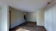 This two-floor space presents a straightforward and functional design with a neutral, understated aesthetic. The main level features dark hardwood flooring that runs throughout, providing a rich contrast against the light grey walls and white ceiling. Natural light filters into the space, casting warm tones across the floor and highlighting the room's open layout. The generous proportions of the main living area offer ample flexibility for a variety of furniture arrangements.

A staircase located toward the rear of the main level connects to the upper floor, featuring dark-toned steps that complement the hardwood flooring below. Overhead, a ceiling-mounted light fixture provides ambient illumination. Baseboard vents are integrated along the walls, indicating a forced-air HVAC system. Electrical outlets are positioned at regular intervals, offering practical utility throughout the space.

The walls are smooth and freshly finished in a clean, neutral tone, giving the space a move-in-ready feel. The overall ambiance is minimal and uncluttered, making it a versatile canvas suited for a range of design preferences and functional needs.