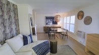 This well-appointed single-floor residence showcases a thoughtfully designed open-plan layout that seamlessly connects the living and dining areas. The living space features a plush white sofa accented with bold navy blue throw pillows and a woven striped area rug, creating a relaxed yet refined atmosphere. A cylindrical metallic side table adds an industrial touch to the seating area.

The dining area is a standout feature, anchored by a sleek rectangular table with a brass-toned frame and surrounded by classic black wishbone-style chairs. Above the table hangs a striking spherical pendant light crafted from natural wood slats, casting warm ambient light throughout the space. A textured stone-faced sideboard lines the back wall beneath a large framed artwork, adding depth and character to the room.

Natural light fills the space generously through plantation-shuttered windows, enhancing the airy, coastal-inspired palette of whites and warm neutrals. Light hardwood flooring runs throughout, contributing to the open and welcoming feel. Crown molding along the ceiling adds a refined architectural detail. The overall ambiance is clean, modern, and inviting — a harmonious blend of contemporary furnishings and classic design elements.