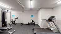 This fitness center is a single-level space designed to provide a well-rounded workout experience within a compact footprint. The room features durable speckled rubber flooring throughout, offering both safety and comfort underfoot during exercise. Overhead, exposed utility conduit runs along the textured ceiling alongside bright fluorescent strip lighting, ensuring the space is consistently and evenly illuminated.

The cardio section is outfitted with multiple Matrix-brand machines, including treadmills, a rowing machine, a stationary spin bike, and an elliptical trainer, providing a variety of cardiovascular workout options. On the strength training side, a Matrix multi-station functional trainer serves as the centerpiece, complemented by a tiered dumbbell rack stocked with a full range of free weights and an adjustable workout bench positioned for use with the cable system.

The walls are painted a clean white, lending the space a bright and functional atmosphere. A hand sanitizer dispenser station is mounted near the entry door, reflecting attention to hygiene. Posted signage throughout the room encourages users to wipe down equipment after each use. Access is provided through a single interior door. The overall design prioritizes practicality and efficient use of space, making the fitness center a functional amenity for everyday use.