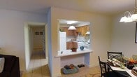 This single-floor space is designed with an open, connected layout that promotes easy flow between living areas. The kitchen is framed by a pass-through opening in the wall, creating visual connectivity with the adjacent dining and living areas while maintaining a defined cooking space. Inside the kitchen, warm honey-toned wood cabinetry lines the walls, offering ample storage, and is complemented by white appliances including a refrigerator, range, and dishwasher. A pendant light hangs above the pass-through counter, providing focused task lighting with a classic touch. The pass-through counter itself doubles as a casual breakfast bar, adding functional versatility to the space. Light beige walls and warm-toned tile flooring run throughout, creating a cohesive and airy atmosphere. A hallway visible from the main area leads further into the residence, with natural light filtering through, suggesting additional rooms beyond. The overall design balances practicality and openness, making the space feel welcoming and efficiently organized for everyday living.