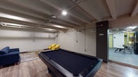 Located at 512 W Ormsby Ave in Louisville, Kentucky, this amenity level offers a thoughtfully arranged recreational and fitness space designed for community use. The space features an exposed beam ceiling with industrial-style piping, lending it a raw, urban aesthetic that is complemented by light wood-style flooring throughout.

At the heart of the space sits a full-size pool table with a sleek black felt surface, complete with a wall-mounted cue rack nearby. Surrounding the billiards table are multiple seating arrangements — a pair of bold yellow accent chairs, a set of brown leather mid-century chairs flanking a side table with a lamp, and a separate lounge nook furnished with a navy blue sofa and dual glass-top gold coffee tables set atop a colorful area rug, providing a relaxed social setting.

An accent wall painted in a muted olive green adds a touch of warmth and color contrast against the painted concrete block walls. Recessed and overhead lighting throughout ensures the space is well-lit.

Adjacent to the main recreation area, a glass-enclosed fitness center is visible, housing a variety of exercise equipment including a cable machine, stationary bike, and weight bench, offering residents a convenient on-site workout option. The overall ambiance is casual, functional, and inviting.