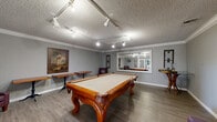 Located at 6016 Sunrise Vista Dr in Citrus Heights, CA, this dedicated billiards room is a well-designed recreational space that combines classic style with functional amenities. The centerpiece is an ornate solid wood pool table with carved legs and a cream felt surface, positioned to allow comfortable play from all sides. The room features durable wood-look luxury vinyl plank flooring throughout, complemented by soft gray walls and crisp white crown molding that lend a polished, finished aesthetic. A textured popcorn ceiling is fitted with adjustable track lighting, ensuring the playing area is well-illuminated at all times. A large wall mirror enhances the sense of space and reflects the room's interior, while decorative wall art, including a prominent tropical-themed painting, adds visual character. Seating is provided by burgundy upholstered armchairs alongside bistro-style side tables with wrought iron bases, creating comfortable spots for spectators. A dartboard mounted near the French glass-paned entry door offers an additional recreational option. The French doors provide a bright, welcoming entry point and connect the room to adjacent areas, allowing natural light to filter in. The overall ambiance is warm, inviting, and purpose-built for leisure and entertainment.