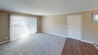 This single-floor residence offers a practical and well-organized layout designed for comfortable everyday living. The living area is generously proportioned, featuring neutral-toned carpet, crown molding, a textured ceiling, and a large sliding glass door dressed with vertical blinds that allows natural light to fill the space. A dedicated entry door provides a clean transition into the main living zone.

The kitchen is efficiently designed, showcasing white cabinetry, a peninsula with a dark countertop that doubles as a breakfast bar, and hardwood-style flooring that extends into the adjacent dining area. The dining space is enhanced by a ceiling fan with integrated lighting and a window with vertical blinds, adding both functionality and natural light. The kitchen connects to a hallway that leads to the remaining rooms, with a laundry area visible along the corridor.

The bedroom is a comfortable, carpeted room with warm neutral walls and a window overlooking outdoor greenery, providing a quiet and private retreat. The bathroom is straightforward in its design, equipped with a tub-shower combination, a toilet, and wood-style flooring, maintaining the cohesive aesthetic found throughout the home.