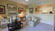 This single-floor residence centers around a warmly decorated living room that immediately establishes a comfortable and classic ambiance. The living room is finished with neutral, earthy-toned walls that complement the soft carpeted flooring, creating a cohesive and inviting atmosphere throughout the space. Framed landscape artwork adorns the walls, adding character and a sense of refinement to the room.

The seating area is anchored by plush, patterned upholstered chairs arranged around a dark wood coffee table, offering a practical yet elegant focal point. A table lamp provides warm accent lighting, enhancing the welcoming feel of the room. Decorative accessories, including ceramic vases and sculptural accents, are thoughtfully placed to add visual interest without overwhelming the space.

A pass-through opening in one wall connects the living room to the kitchen, which features light wood cabinetry and white appliances visible from the main living area. This open connection allows natural sightlines between the two spaces, contributing to an airy, functional layout. A hallway visible from the living area suggests additional rooms beyond, completing this well-organized single-floor layout.