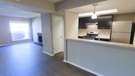This single-floor residence showcases an open-concept layout that seamlessly connects the kitchen, dining, and living areas. The kitchen is defined by dark espresso cabinetry that contrasts with light granite-style countertops, a stainless steel electric range, and an under-cabinet range hood. A peninsula with a breakfast bar separates the kitchen from the adjacent spaces, offering both functionality and a casual dining option. A pendant light fixture hangs above the peninsula, adding a focused decorative touch. Dark hardwood flooring runs consistently throughout, grounding the space with warmth and continuity. The living area, visible through an open passageway, features a striking floor-to-ceiling stone fireplace that serves as a focal point, adding character and a cozy ambiance. The neutral gray walls throughout provide a clean, versatile backdrop. A white interior door leads to an additional area of the home, hinting at further private spaces beyond. Natural light filters in from windows in the living area, brightening the overall atmosphere of this well-connected, functional layout.