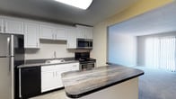 Located at 1445 Bodwell in Manchester, New Hampshire, this single-floor residence offers a well-designed layout with a cohesive, neutral aesthetic throughout. The kitchen is a standout feature, equipped with stainless steel appliances including a refrigerator, electric range, over-the-range microwave, and dishwasher. Crisp white cabinetry provides ample storage, while the dark, marbled countertops add a striking contrast. A breakfast bar extends into the adjacent living area, creating a seamless open connection between the two spaces. The living area is carpeted in a soft gray tone and features a sliding glass door fitted with vertical blinds, allowing natural light to filter in while providing access to the outdoors. A wall-mounted air conditioning unit and baseboard heating ensure year-round comfort. The bedrooms are comfortably sized with plush carpeting, neutral walls, and textured ceilings. One bedroom includes mirrored sliding closet doors, adding both storage and a sense of depth to the room, while another features a window with an outdoor view. The bathroom is neatly appointed with white cabinetry, a dark countertop vanity, a large mirror, a shower-tub combination, and wood-look flooring, completing the unit with practical, clean finishes throughout.