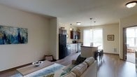This single-floor residence offers an open-concept layout that seamlessly connects the living, dining, and kitchen areas into one cohesive and functional space. The kitchen is appointed with rich dark cabinetry, black appliances including a full-size refrigerator and microwave, and a deep sink positioned beneath a large window that fills the space with an abundance of natural light. Pendant lighting fixtures hang above the kitchen and breakfast bar, adding warmth and style to the area. The breakfast bar, fitted with bar-height stools, provides a casual dining option and serves as a natural divider between the kitchen and the rest of the open living space. Hardwood-style flooring runs throughout, lending a clean, modern aesthetic and a sense of continuity across the floor plan. The dining area accommodates a full table and sits adjacent to the living room, which is furnished with a comfortable sofa accented with decorative pillows. Neutral wall tones in soft beige create a warm, inviting ambiance throughout. The large window in the kitchen area allows for generous natural light to penetrate deep into the space, enhancing its open and airy character. Overall, this space is well-designed for both everyday living and casual entertaining.