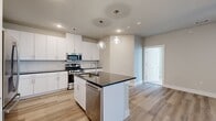 This well-appointed 3-bedroom, 2-bathroom single-story home offers a thoughtfully designed open-concept layout. The kitchen serves as a focal point, featuring crisp white cabinetry, dark granite countertops, a subway tile backsplash, and a full suite of stainless steel appliances including a gas range, microwave, and dishwasher. A central island with an integrated sink and matching granite top provides additional prep space and seating. Globe pendant lights add a modern touch overhead. Light wood-style plank flooring flows through the kitchen and living areas, while the living room benefits from natural light through a glass-paneled entry door and sidelights, complemented by a ceiling fan. One bedroom features a built-in desk with dark granite surface and white upper cabinetry — ideal as a home office. The carpeted bedrooms offer ceiling fans and windows with outdoor views. Each bedroom is supported by well-sized closets with built-in shelving and hanging rods. The bathrooms are finished with white vanity cabinetry, dark granite counters, and a glass-enclosed walk-in shower adorned with marble-style tile. Neutral warm tones and clean finishes throughout create a bright, cohesive ambiance.