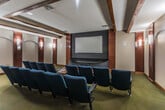 Located in Cordova, TN, this private movie theater delivers an immersive cinematic experience within a thoughtfully designed single-floor layout. The main screening room is the centerpiece of the space, anchored by a large projection screen mounted against a bold black accent wall, complemented by a curved AV console beneath it. Rows of plush, navy blue theater-style seats with armrests and cup holders are arranged to provide clear sightlines to the screen, offering a true cinema atmosphere.

The walls are dressed in warm wood paneling with decorative arched niches, each flanked by elegant wall sconces that cast a soft, ambient glow throughout the room. Exposed wooden ceiling beams add a classic architectural character, while the textured ceiling and deep-toned carpet flooring contribute to the space's acoustic and visual warmth.

At the rear of the screening room, a built-in refreshment nook features cabinetry and counter space with a microwave, providing convenient in-theater amenities. Multiple doors lead to a closet and a utility room, ensuring practical functionality is seamlessly integrated. Exit signage and a fire extinguisher reflect attention to safety. Overall, this space combines refined design details with purpose-built features to create an intimate and well-appointed private theater experience.
