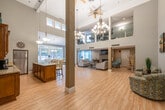 This expansive single-floor space is defined by its dramatic soaring ceilings and open-concept layout, creating an airy, welcoming atmosphere throughout. Light-toned hardwood flooring flows seamlessly across the entire space, unifying its distinct functional areas.

The kitchen is well-appointed with rich warm-toned cabinetry, granite countertops, stainless steel appliances including a full-size refrigerator and dishwasher, and a central island with bar seating for three. Pendant lighting overhead adds a refined touch. Upper cabinets with glass-front panels provide both storage and visual interest.

Adjacent to the kitchen, a bright dining area features large picture windows that frame views of an outdoor patio and surrounding greenery. A glass-top round dining table beneath a chandelier anchors the space, filled with natural light.

The lounge area offers two sofas, a stone-surround fireplace with a wall-mounted television, and teal accent drapes. A graceful spiral staircase nearby adds an architectural focal point. A secondary seating nook displays patterned accent chairs beneath large abstract wall art, complemented by the towering ceilings above.

Ceiling fans and chandeliers throughout ensure both comfort and ambiance, making this a versatile, thoughtfully designed communal space.