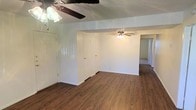 This single-story space presents a straightforward, functional layout with a clean and neutral aesthetic. The main living area is finished with warm-toned hardwood-style flooring that extends throughout, creating a cohesive and easy-to-maintain surface underfoot. White walls provide a bright, neutral backdrop, while ceiling fans with integrated light fixtures are installed in both the main area and an adjoining room, ensuring air circulation and ambient lighting throughout.

The textured ceiling adds a classic residential character to the space. Multiple doorways are visible from the main living area, suggesting a practical floor plan with clearly defined rooms. Two interior doors are positioned along one wall, potentially leading to closets or additional rooms, while a hallway extends further into the unit, hinting at additional living or sleeping spaces beyond the main area.

The open sightlines between rooms contribute to a sense of connectivity throughout the layout. The overall ambiance is clean, neutral, and move-in ready, offering a blank canvas that accommodates a variety of furnishing styles and personal preferences.