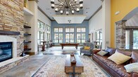 Located at 5350 Old Dowlen Rd in Beaumont, Texas, this single-floor community living room is a thoughtfully designed gathering space that blends elegance with comfort. Soaring ceilings with a coffered tray detail draw the eye upward, while a dramatic multi-arm chandelier serves as a striking centerpiece overhead. The space is divided into distinct social zones, beginning with a cozy seating area anchored by a floor-to-ceiling stacked stone fireplace with a rustic wood mantel and flanking built-in shelves — a true architectural focal point. An adjoining billiards area features a full-sized pool table positioned beneath pendant lighting and framed by an expansive bank of gold-trimmed windows, flooding the room with natural light and offering views of the outdoor pool area. A vibrant yellow accent wall with a stone-framed arched passageway leads to an adjoining area, adding a bold pop of color and visual depth. Durable tile and hardwood-style flooring transition seamlessly between zones, while layered area rugs in soft, eclectic patterns add warmth. The furnishings — leather sofas, striped armchairs, and bistro-style high-top tables — create an inviting, versatile environment suited for both relaxation and socializing.