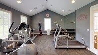 This single-floor fitness center is thoughtfully designed to support a comprehensive workout experience. The space features vaulted ceilings that lend an open, airy feel, complemented by recessed lighting and a neutral gray color palette that creates a focused, energizing environment. Large windows fitted with white blinds line the walls, flooding the room with natural light and offering views of the surrounding greenery outdoors.

The facility is outfitted with a diverse selection of cardio equipment, including elliptical trainers, a stationary bike, and treadmills, ensuring options for all fitness levels. Strength training is well represented with a power rack complete with a bench and weight plates, a cable machine, and a dedicated free weights area featuring a full dumbbell rack and wall-length mirror — ideal for form checks during training. A freestanding punching bag adds variety for those seeking a more dynamic workout.

Durable, patterned carpet tiles cover the floors throughout, providing both comfort and practicality. The space is accessible through elegant French doors with glass panels. A wall-mounted television and motivational wall decals contribute to the overall atmosphere. Whether focusing on cardio endurance, strength building, or flexibility, this fitness center provides a well-rounded environment for achieving a variety of health and wellness goals.