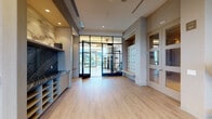 This contemporary fitness center is designed to offer a premium workout experience across a thoughtfully organized single-floor layout. The entryway welcomes users with sleek wood-look flooring, built-in cabinetry with cubbies, a dark stone-accented counter, and a bank of individual lockers — providing practical storage before stepping into the main exercise areas through steel-framed glass doors.

The main gym floor is divided into distinct functional zones. The cardio area is lined with treadmills, stair climbers, ellipticals, and stationary bikes, all positioned to take full advantage of the expansive floor-to-ceiling glass walls that frame sweeping panoramic city views. Natural light floods the space throughout the day, creating an open and energizing atmosphere.

A dedicated strength and free weights zone features adjustable benches, a squat rack, dumbbells, medicine balls, and cable machines. Full-length mirrors along one wall amplify the sense of space and allow for proper form monitoring. The free weights area is carpeted in durable speckled flooring consistent throughout the gym.

Overhead, recessed linear lighting provides even, well-distributed illumination. The overall design palette — neutral grays, warm wood tones, and muted finishes — gives the facility a polished, upscale aesthetic suited to a high-end amenity space.
