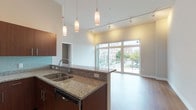 This contemporary single-floor residence is thoughtfully designed with an open-concept layout that seamlessly connects the kitchen, dining, and living areas. The kitchen serves as a central hub, featuring rich dark wood cabinetry, granite countertops, a glass tile backsplash, and a full suite of stainless steel appliances including a French door refrigerator, range, and built-in microwave. A peninsula with a double stainless steel sink and pendant lighting adds both functionality and style, while track lighting overhead ensures the space is well illuminated throughout.

The living area is anchored by expansive floor-to-ceiling windows and a sliding glass door that flood the room with natural light and frame views of the surrounding urban streetscape. Warm hardwood-style flooring flows through the main living spaces, creating a cohesive and inviting atmosphere.

The bedroom offers plush carpeting, a closet with built-in shelving, and a window that brings in soft, diffused light. Two bathrooms are finished with large-format tile flooring, granite-topped dark wood vanities, and clean, neutral tones. One bathroom includes a glass-enclosed walk-in shower with a ceiling-mounted fixture. A dedicated laundry room with a full-size washer and dryer adds everyday convenience to this well-appointed residence.