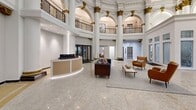Located at 265 College St in New Haven, CT, this stunning single-floor lobby showcases a masterful beaux-arts architectural design that commands attention from every angle. The space is anchored by a dramatic rotunda framed by towering Corinthian columns adorned with intricate gold-leaf capitals, lending a sense of grandeur and historic significance. Soaring ceilings draw the eye upward to an ornate upper-level balcony wrapped in elegant wrought-iron railings with decorative geometric patterns, accessible via the surrounding mezzanine level. The floor is finished in polished terrazzo tile with mosaic detailing, reflecting light beautifully throughout the expansive open area. A sleek, curved reception desk with warm gold and neutral tones anchors one side of the room, offering a modern contrast to the classical architecture. A tastefully arranged seating area with a navy blue sofa, leather armchairs, and a low coffee table provides a welcoming focal point at the center of the space. Glass-paneled interior partitions and doorways allow views into adjacent utility and fitness areas. The overall ambiance harmoniously blends historic architectural opulence with contemporary furnishings, creating an impressive and functional lobby experience.