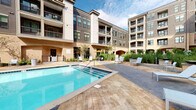 Located at 4206 Simcoe Ln in Louisville, Kentucky, this outdoor amenity space is part of a contemporary multi-story residential complex defined by a striking combination of brick, dark horizontal siding, and large windows. The centerpiece of the outdoor area is a generously sized swimming pool with clear blue water, featuring a gradual entry point and accessible steps, surrounded by a stamped concrete deck that mimics the appearance of natural wood and stone.

Encircling the pool, the sun deck is furnished with modern sling-style lounge chairs and side tables, providing ample space for relaxation. Raised planter beds line portions of the perimeter, filled with ornamental grasses, flowering trees, and structured evergreen shrubs that introduce lush greenery and visual interest throughout the courtyard. Iron fencing defines the boundaries of the space while maintaining an open, airy feel.

The building's architecture frames the courtyard on multiple sides, with private balconies overlooking the pool area from upper floors. Covered archway entries and additional seating areas with shade umbrellas complement the space. The overall ambiance is that of an upscale, resort-inspired outdoor retreat, thoughtfully landscaped and designed for both active recreation and leisurely outdoor enjoyment.