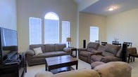 This single-floor home makes a strong impression with its open and airy living space, designed to maximize comfort and natural light. The living room serves as the centerpiece of the home, boasting soaring vaulted ceilings that lend a grand sense of scale to the space. A striking arched window crowns the wall alongside multiple large windows dressed with horizontal blinds, flooding the room with abundant natural light and creating a warm, inviting atmosphere.

The living area is generously proportioned, offering ample room for relaxed seating arrangements. Soft, neutral-toned carpeting underfoot complements the warm beige walls, contributing to the overall sense of calm throughout. The layout flows naturally, with a glimpse of a dining area visible in the background, suggesting a well-connected and functional floor plan.

The space is finished with smooth walls and clean architectural lines, reflecting a classic residential design sensibility. The combination of vaulted ceilings, well-placed windows, and an open layout makes this home feel expansive and bright throughout the day. Whether used for entertaining or everyday living, this single-floor property offers a comfortable and thoughtfully designed environment that balances spaciousness with warmth.