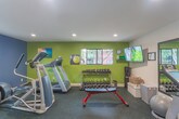 Located at Glenwood Circle in Monterey, California, this fitness center is a well-appointed, single-floor facility designed to support a comprehensive workout routine. The space is divided into distinct exercise zones, each thoughtfully equipped and finished.

The cardio area features a treadmill and an elliptical trainer, positioned beneath recessed ceiling lights and framed by bold navy blue and olive green accent walls. Windows along the perimeter allow natural light to filter in, offering views of the surrounding greenery and creating an energizing atmosphere. Motivational artwork adds a personal touch to the space.

On the opposite side of the room, a row of full-length mirrors mounted in black frames visually expand the space while allowing users to monitor their form. A stationary spin bike, a stability ball, and a flat-screen TV mounted on the wall complete this section, alongside a hand sanitizer station for convenience.

A multi-functional cable and pulley strength machine anchors the strength training zone, complemented by a wall-mounted resistance band station. A dedicated free weights area features a well-stocked dumbbell rack alongside a weight bench, catering to a range of strength training needs.

The flooring throughout is durable rubber matting, ideal for high-impact activity. Direct access to an outdoor patio area is available through a glass-panel door, seamlessly connecting the indoor fitness space with the exterior environment.